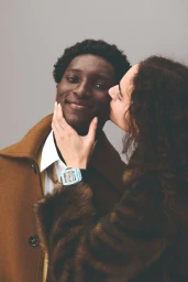 Close-up of a smiling black man wearing a brown coat and white shirt, with a woman leaning in to touch his face against a grey studio background