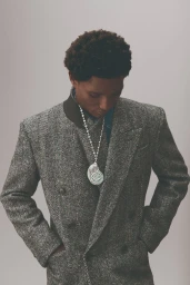 A Black man with short dark hair wearing a grey tweed double-breasted coat and a large pendant necklace against a plain grey background