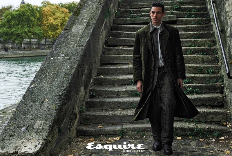 A man in a long green coat walks down stone steps next to a river in an outdoor fashion editorial, shot at eye-level.
