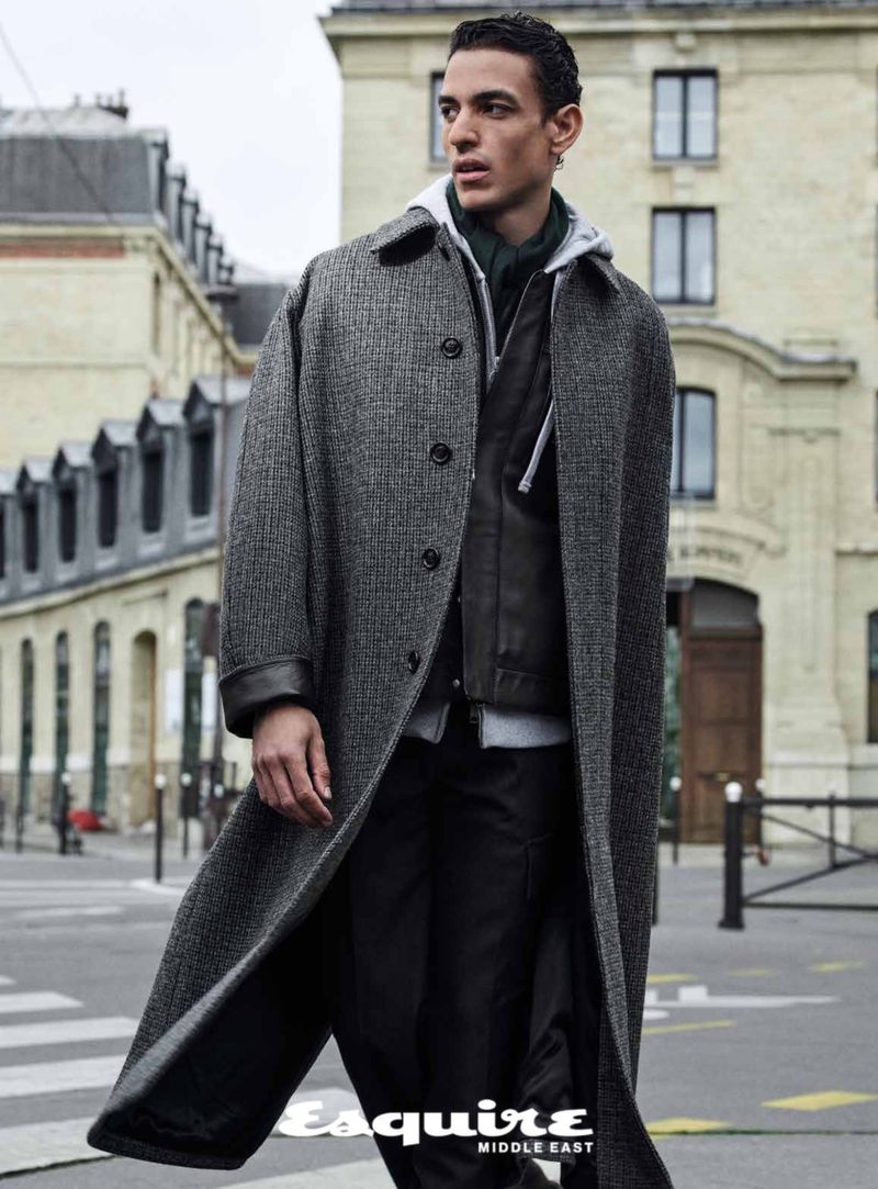 A fashion editorial shot of a male model wearing an overcoat, captured outdoors against a cityscape background, for Esquire Middle East.