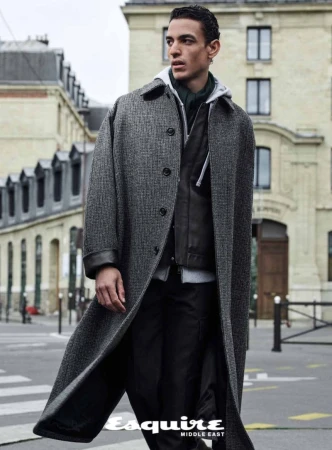 A fashion editorial shot of a male model wearing an overcoat, captured outdoors against a cityscape background, for Esquire Middle East.