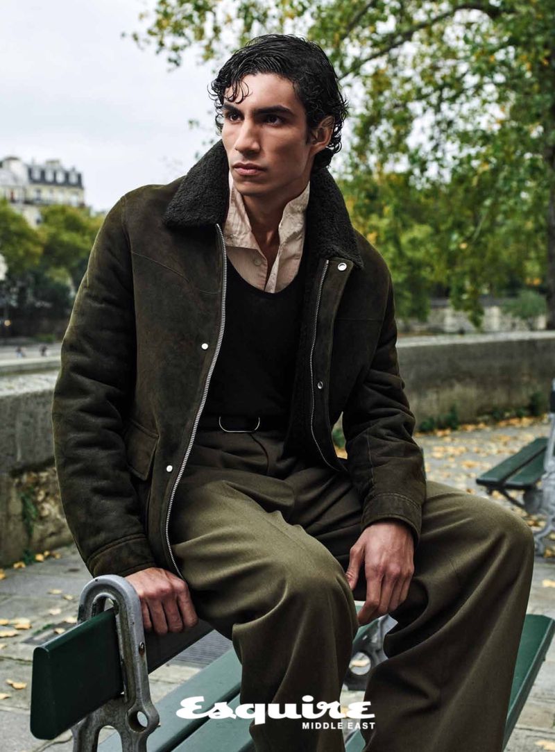 A young adult man poses in a shearling jacket and olive-green pants on a park bench with an urban background in a fashion editorial.