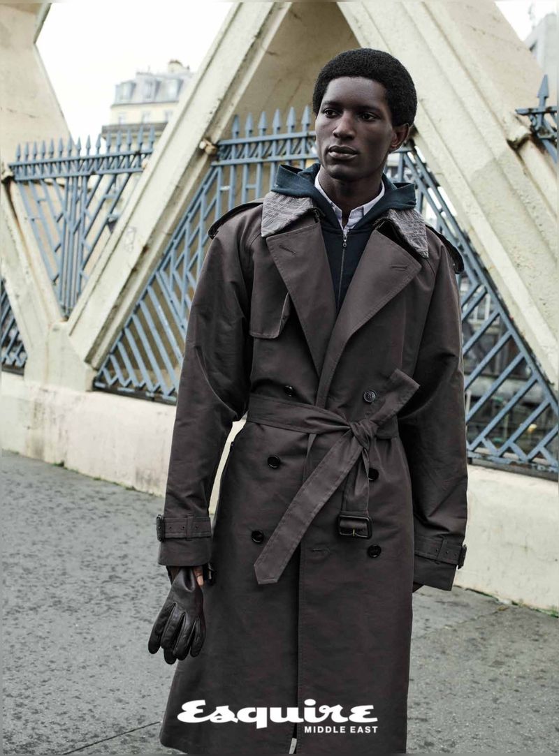 Black man wearing a dark belted trench coat standing outdoors near architectural stone structures and metal fencing.