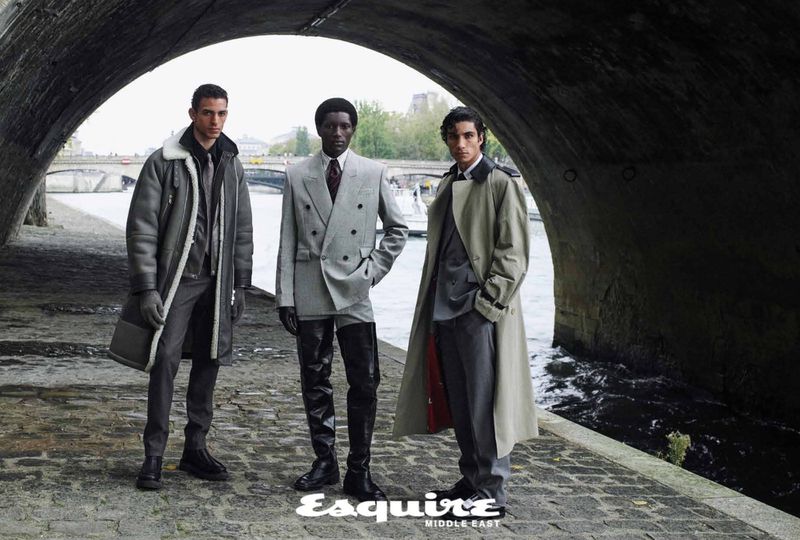 Three male models stand under a stone bridge in a fashion editorial. One wears a shearling coat, another a gray suit, and the last a trench coat.
