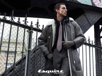 Man wearing a shearling-lined grey coat and tie standing beside an ornate iron fence outdoors.