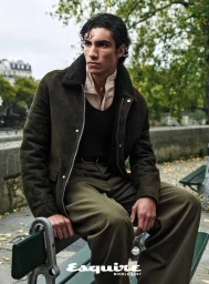 Man wearing an olive green shearling collar jacket and trousers seated on a park bench outdoors