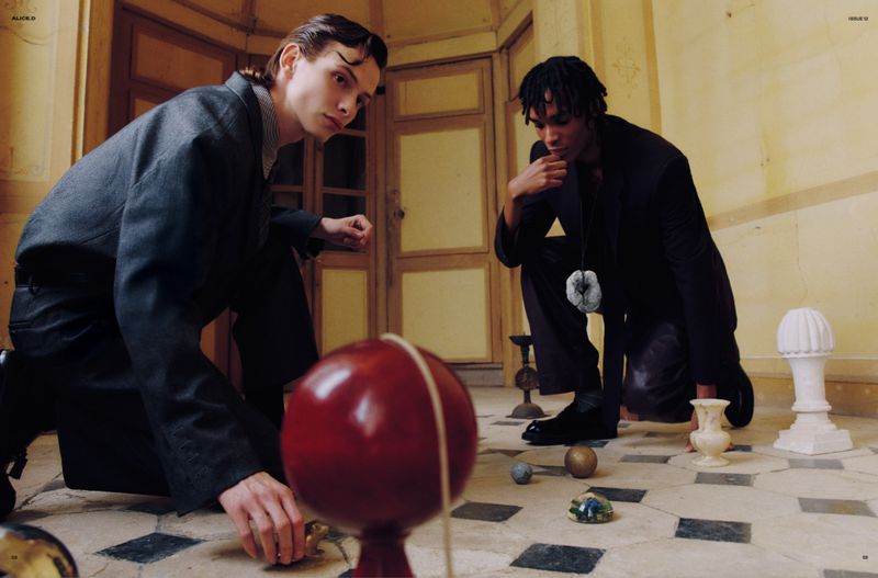 Two male models pose indoors with spheres; one crouching, one kneeling, wearing dark suits against a cream and black tiled backdrop.