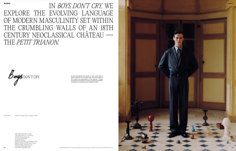 A man stands in a suit, indoors with geometric floor tiles and various small decorative objects, posing formally in a magazine editorial.