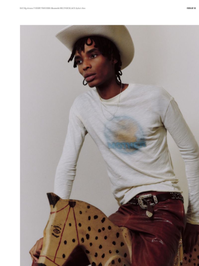 Young adult in white cowboy hat and long-sleeve shirt sits astride a vintage toy horse in a studio setting.