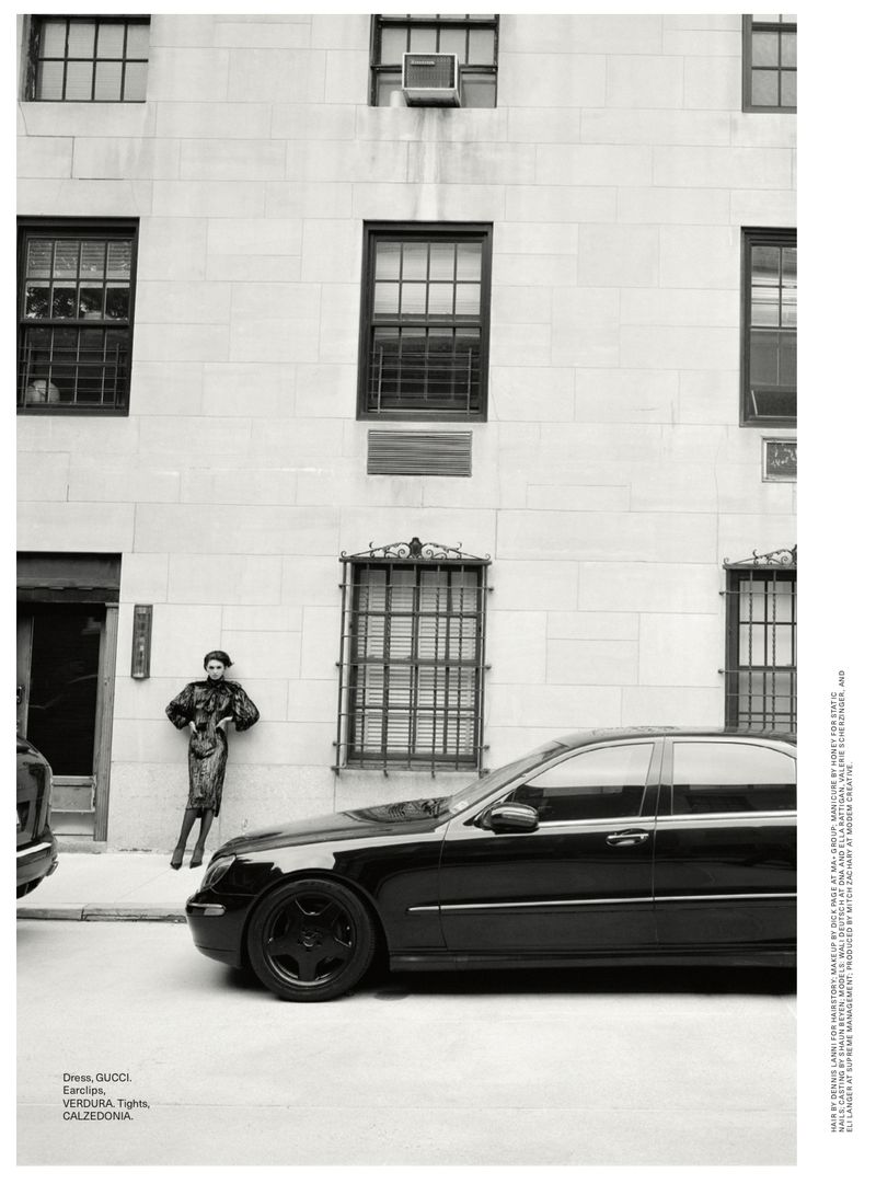 Monochrome outdoor shot of an adult female model in a patterned dress, leaning against a stone building next to a black luxury sedan.