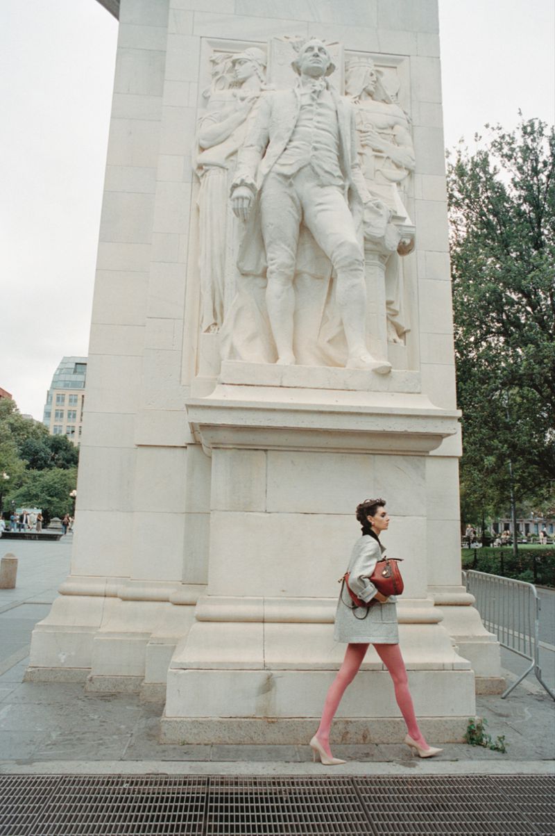 A woman with a satchel bag walks past the Washington Square Arch, wearing a skirt suit with pink tights and heels in an outdoor setting.