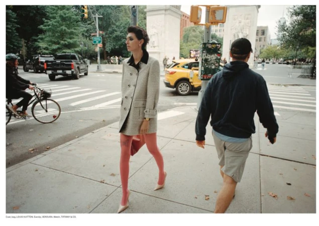An adult female in a grey coat and pink tights walks on a sidewalk in a city with cars, pedestrians, and buildings in the background.