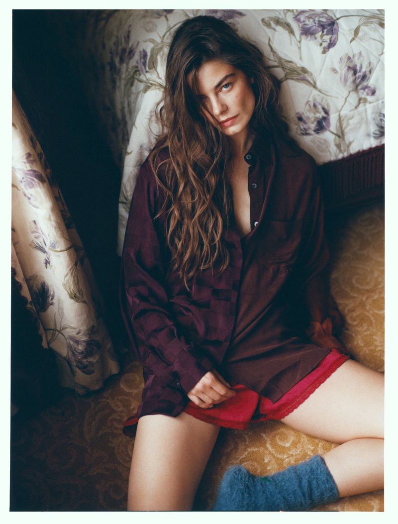 Woman with long wavy hair poses in an unbuttoned maroon shirt and red shorts while reclining on a patterned surface with soft lighting.