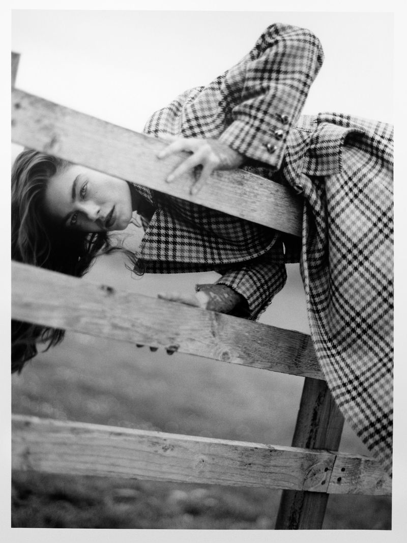 Black-and-white fashion shot of a woman in a plaid coat leaning over a wooden fence with a neutral expression on an overcast day.