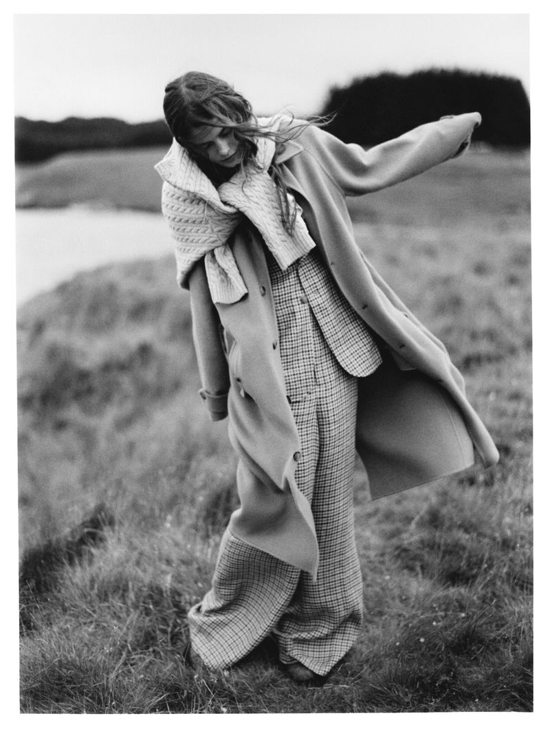 A woman in a long coat and patterned pants stands in a field of grass near a body of water under an overcast sky in a black and white editorial.