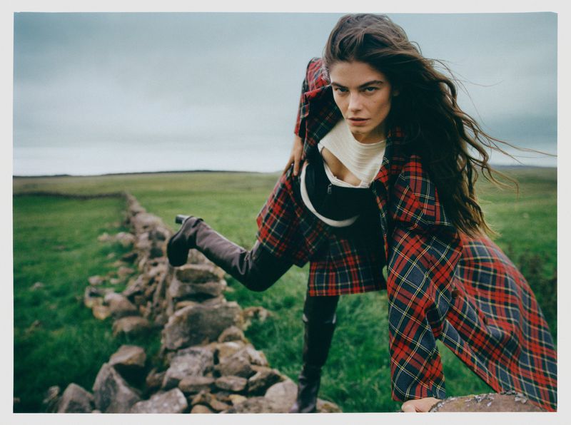 Brunette woman in red plaid coat and black boots poses over stone wall in grassy field under cloudy sky in outdoor fashion shot.