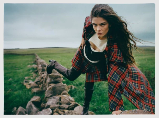 Brunette woman in red plaid coat and black boots poses over stone wall in grassy field under cloudy sky in outdoor fashion shot.