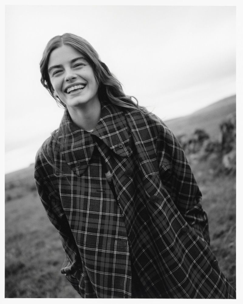Smiling woman in a plaid coat stands outside in a black and white photo, with a field and cloudy sky in the blurred background.