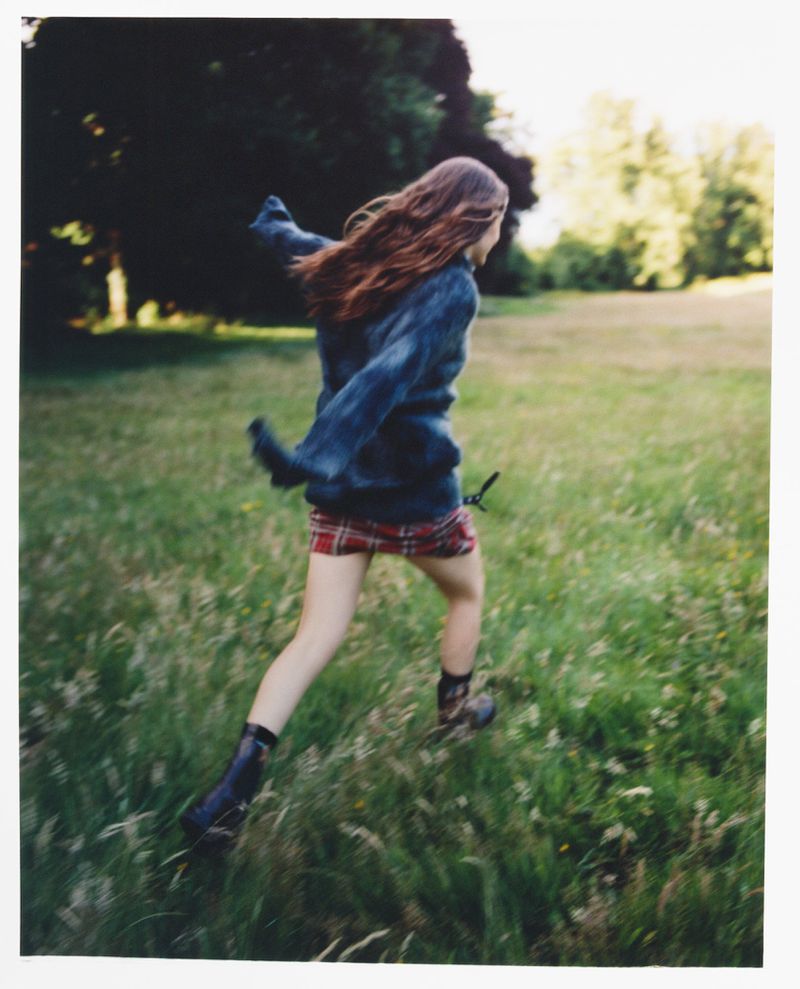 A person is seen from behind running through a grassy field on a sunny day, wearing a blue sweater and red plaid skirt.