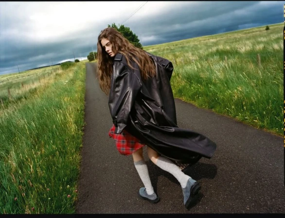 A long-haired woman in a black coat and red plaid skirt glances back while walking on a road through a grassy field under a cloudy sky.
