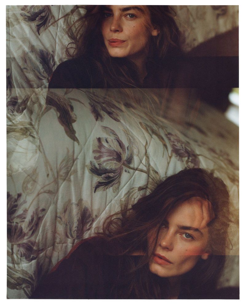 Woman with long brown hair poses on a floral quilt in a close-up portrait. The image has soft, diffused lighting and a shallow depth of field.