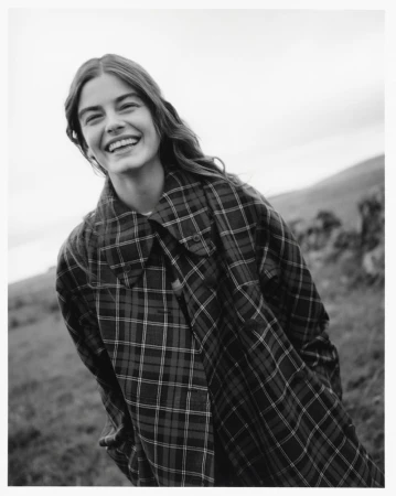 Smiling female model in a plaid coat outdoors against a natural landscape background