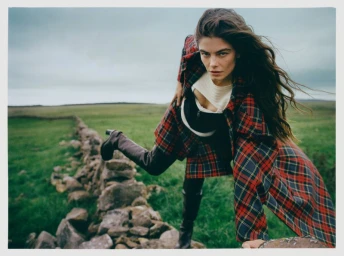 Model in red plaid coat leaning over a low stone wall in a grassy landscape under an overcast sky.