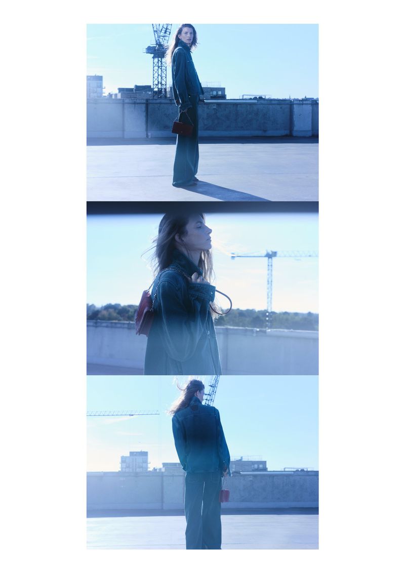 A woman in a jacket and wide-leg trousers poses on a rooftop on a sunny day, accessorized with a small purse and a backpack.