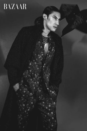 Monochrome fashion shot of a male model wearing a patterned suit and dark overcoat, posing in a studio against a grey background.