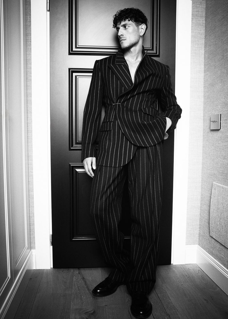 Man wearing a dark pinstripe suit standing against a dark paneled door in an interior hallway.