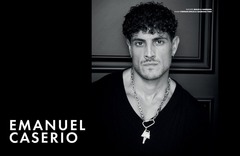 Monochrome portrait of a man with curly hair wearing a dark sweater and silver chain with a doorframe in the background.