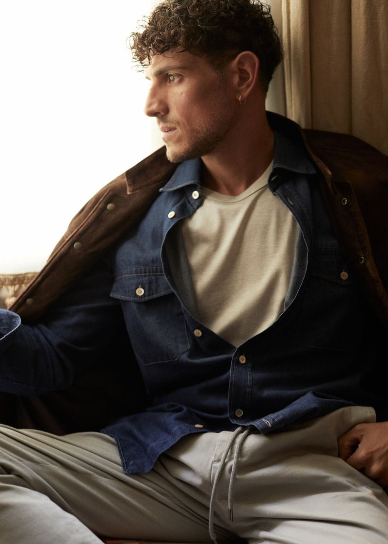 Portrait of a curly-haired adult man in a blue denim shirt and grey sweatpants sitting indoors looking away from the camera.