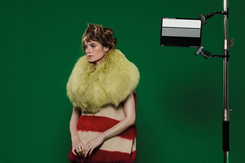 Adult female posing in a studio with a green screen backdrop, wearing a furry yellow collar and a striped red and cream dress.