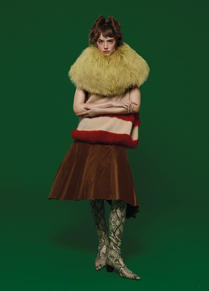 Full-length portrait of a person in a beige and red striped top, brown skirt, high patterned boots, and fur collar against a green background.