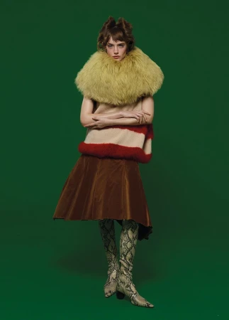 Full-length portrait of a person in a beige and red striped top, brown skirt, high patterned boots, and fur collar against a green background.