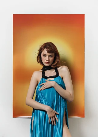 A fashionable woman poses in a blue dress against a gradient orange backdrop, creating a striking editorial image in a studio setting.