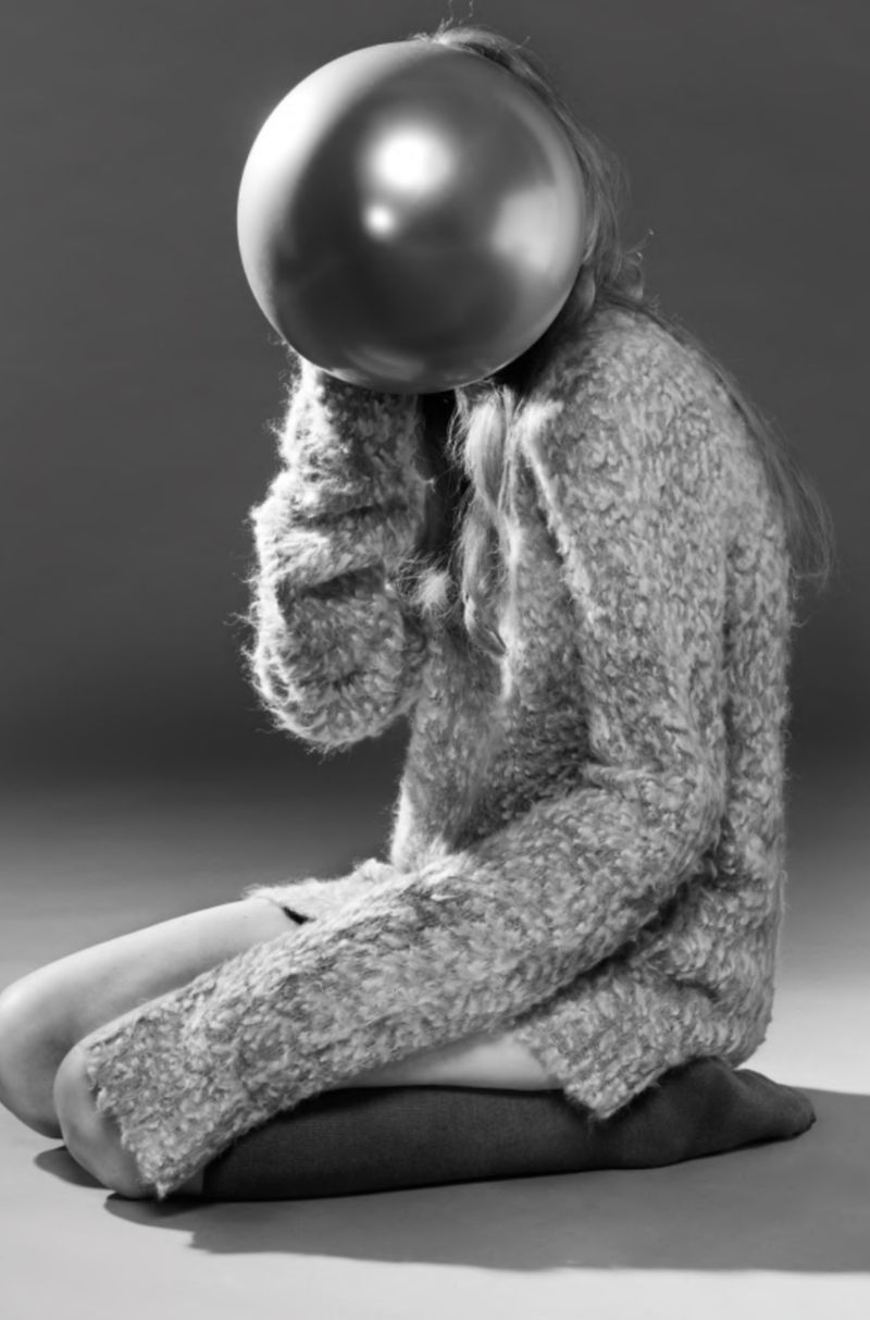 A young woman sits cross-legged on the floor. A metallic balloon obscures her face, creating a surreal and monochrome portrait.