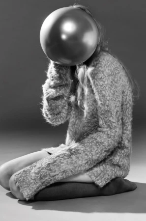 A young woman sits cross-legged on the floor. A metallic balloon obscures her face, creating a surreal and monochrome portrait.