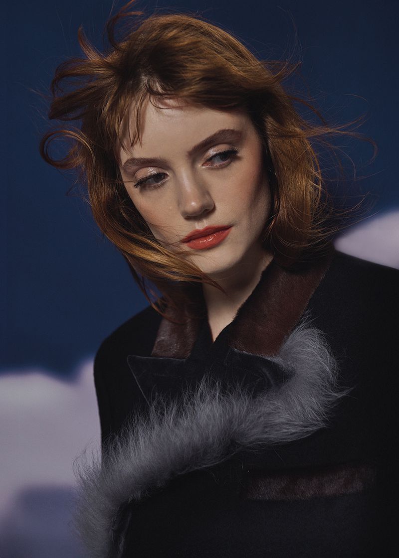 Portrait of a red-haired woman with fair skin wearing a black coat against a cloudy blue background, fashion, beauty, studio lighting.