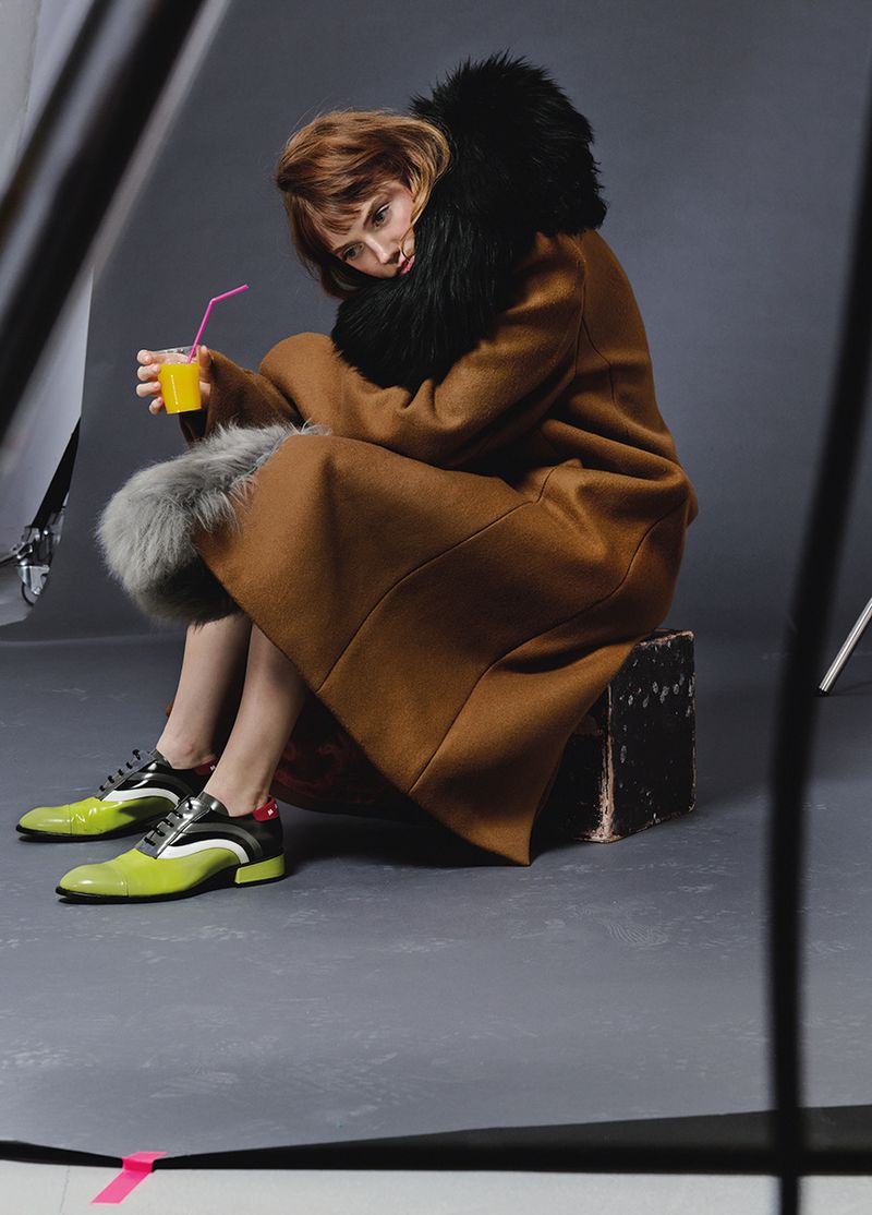 Woman in a brown fur-trimmed coat sits with a beverage and straw, posing in a studio with gray backdrop and artificial lighting.