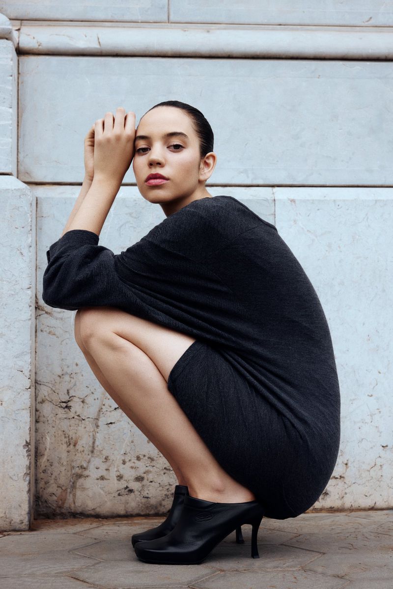 Studio shot of a young woman in a grey dress, squatting, with her hands near her face, and wearing black high-heeled shoes.