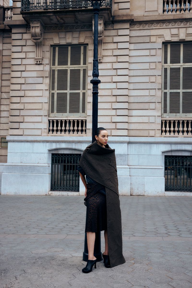 Fashionable woman stands in front of a light post and a large building, wearing a brown shawl and black dress, striking a pose.
