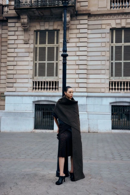 Fashionable woman stands in front of a light post and a large building, wearing a brown shawl and black dress, striking a pose.