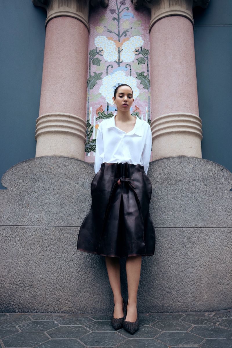 Young woman in fashionable outfit stands outdoors against wall with columns and mosaic art. Brown skirt, white top, neutral color palette.