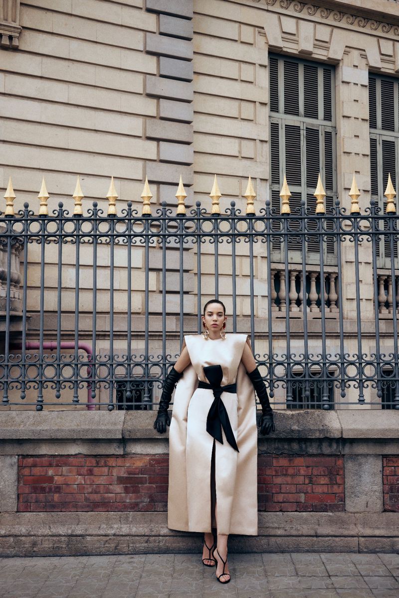 A woman in a cream-colored dress and long black gloves poses outdoors in front of a black iron fence and historic building.