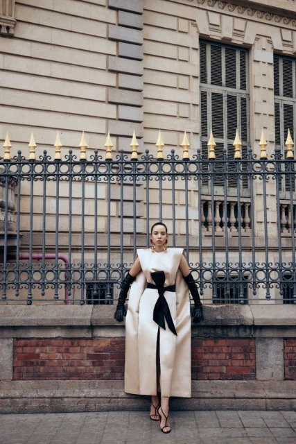 A woman in a cream-colored dress and long black gloves poses outdoors in front of a black iron fence and historic building.