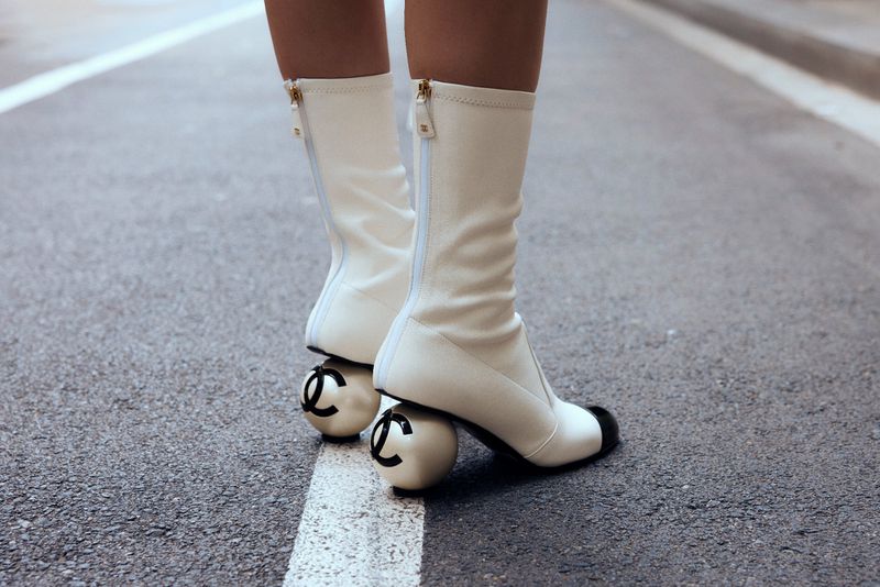 Close-up of white ankle boots with black toe and spherical heel featuring the black Chanel logo, posed on an asphalt road with a white line.