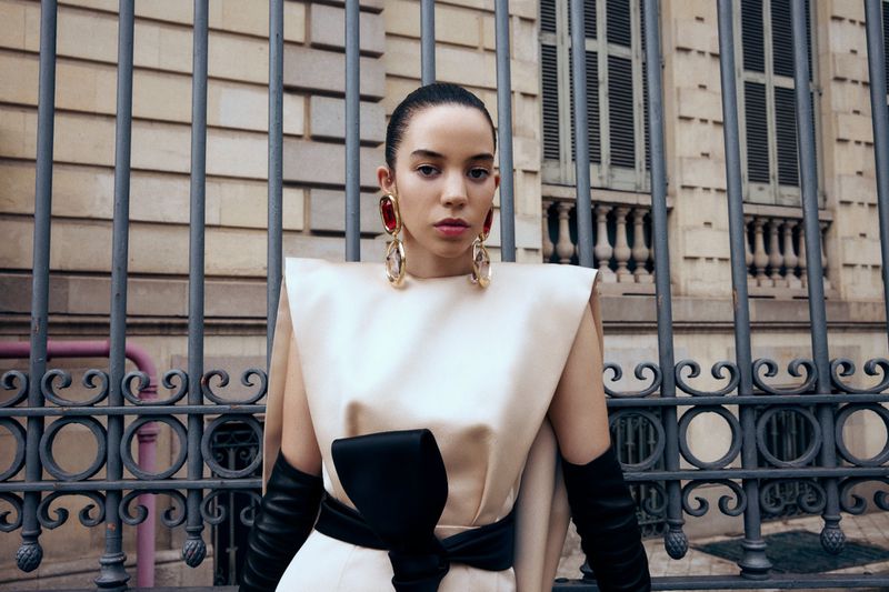 Elegant woman poses in front of an iron gate wearing a cream dress with a black bow, long black gloves, and large gold and ruby earrings.