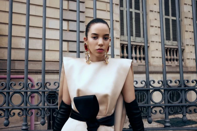 Elegant woman poses in front of an iron gate wearing a cream dress with a black bow, long black gloves, and large gold and ruby earrings.