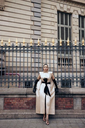 Model Agustina Yanque wearing a cream satin gown with a large black bow belt and long black gloves, standing in front of an iron fence.